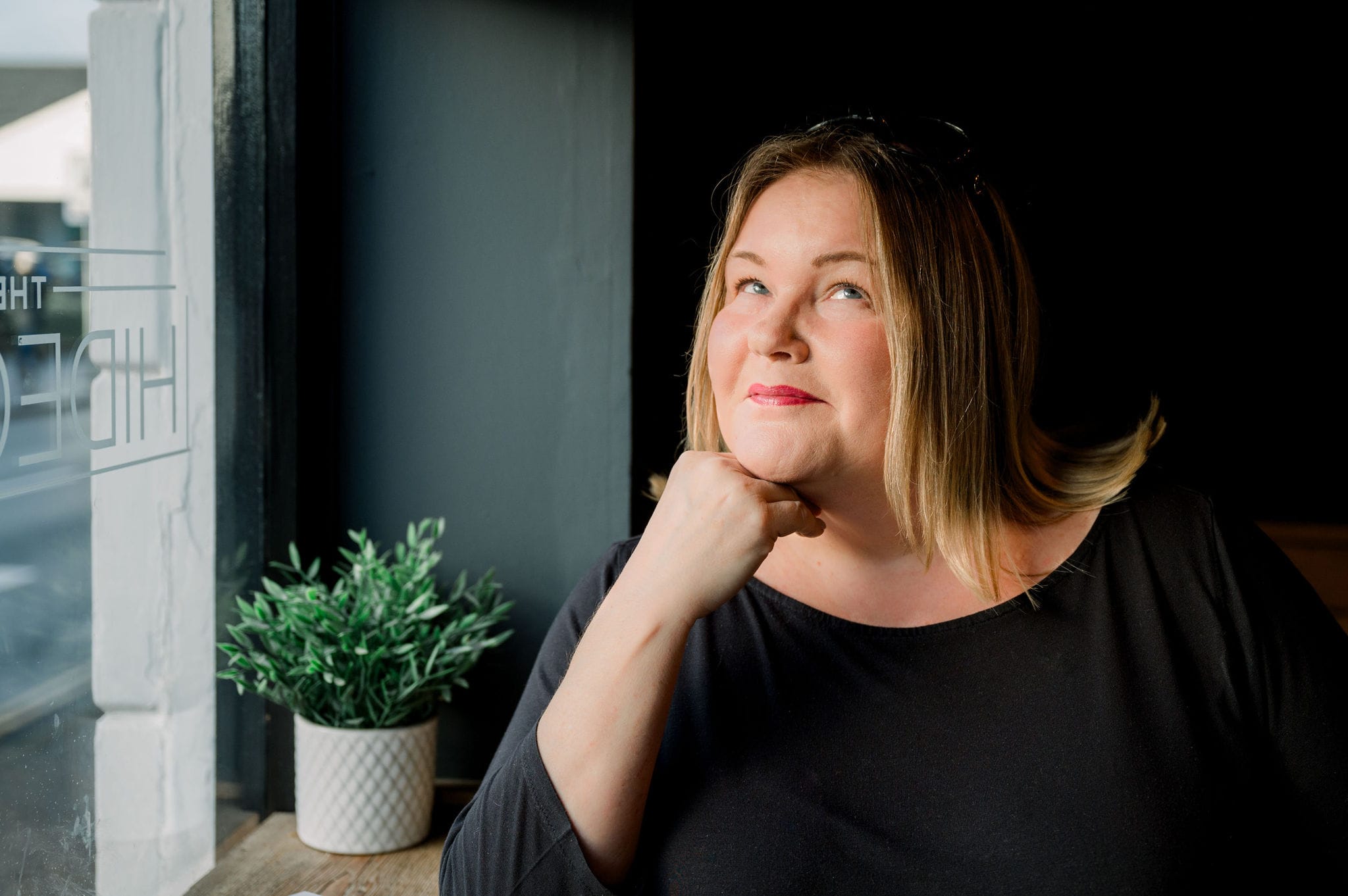 A woman with blonde hair, wearing a black top, sits by a window with her chin resting on her hand, gazing upwards thoughtfully. A small potted plant is on the windowsill beside her.